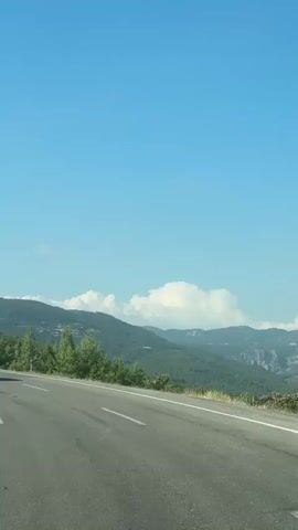 The video shows a highway with two cars. One car is black, and the other is red. The black car is in front of the red car. The highway is surrounded by trees and mountains. The sky is clear and blue.