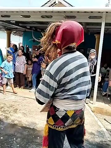 The video shows a street performance, likely a cultural dance or parade, taking place outdoors on a sunny day. The foreground features a performer dressed in a striped shirt, patterned pants, and a red head covering, with elaborate face paint and a long, flowing mane-like wig. This performer is facing away from the camera and appears to be in motion. To the left, another performer is visible, also in elaborate costume, with a large, feathered headdress and a mask-like face. This performer is facing the camera, and the words "Gondorong" are visible on their costume. In the background, a crowd of spectators, including men, women, and children, are gathered to watch. Some are standing, while others are sitting. They are dressed in casual clothing. Behind the performers and spectators, a building with a white roof and pillars is visible, suggesting a residential or community area. There are also some buckets and debris on the ground, indicating the performance is taking place on a paved or dirt surface. The overall atmosphere appears festive and engaging.