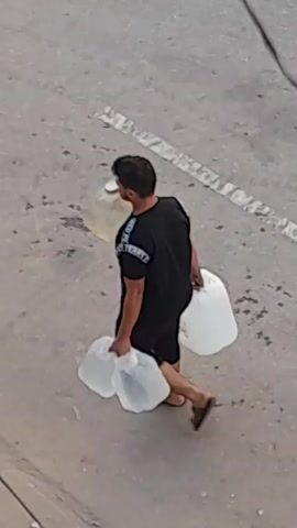 A person is walking while carrying four large plastic jugs, three of which appear to be full of a liquid. The person is wearing a black t-shirt and black shorts with flip-flops. The person is walking on a paved surface, possibly a road or sidewalk. There are some debris on the ground. There is a utility pole in the background. The image is slightly blurry, and the lighting suggests it is daytime.