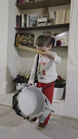 Here's a description of the video:

A young boy, approximately 4 years old, stands indoors and plays a drum. He is wearing a long-sleeved white shirt with a Christmas-themed graphic on the front and red pants. He has short brown hair. The drum is strapped across his chest, and he's holding a drumstick in his right hand, using it to hit the drum. There’s a white and gray patterned towel tucked under the drum.

Behind the boy is a built-in wall shelf unit. The shelves contain books, picture frames, potted plants, and various decorative items. The wall appears to be white with some texture. The flooring is light-colored tile.