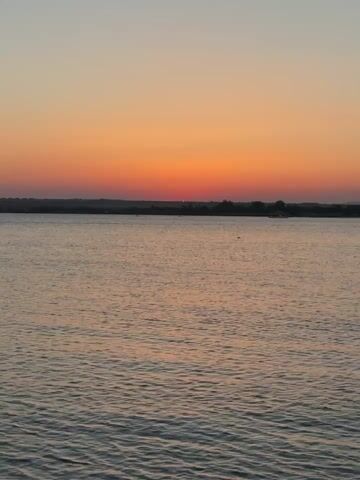 The video displays a body of water with a horizon line in the distance. Above the horizon, the sky transitions from a pale yellow at the top to a vibrant orange and then a deep red near the horizon. The water has ripples, suggesting a slight breeze or current. The horizon appears to be a treeline or a low-lying landmass. There are no people or distinct objects visible on the water or land. The overall scene captures a sunset or sunrise over a calm body of water.