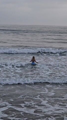 A woman with long blonde hair, wearing a blue swimsuit and a red shirt, stands in the ocean as waves break around her. The sky is overcast, creating a muted, greyish light. The water is choppy, with white foam visible on the crests of the waves. The woman is seen from behind and appears to be facing the horizon. After a moment, she wades out of frame.