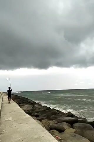 A video depicts a man fishing on a concrete walkway beside the sea under a cloudy sky. The man is seen standing at the edge of the walkway, holding a fishing rod above his head. The sea is choppy with small waves breaking along the rocky shoreline. Large, dark clouds dominate the sky, suggesting an overcast or stormy day. The walkway extends into the distance, bordered by large rocks acting as a breakwater.