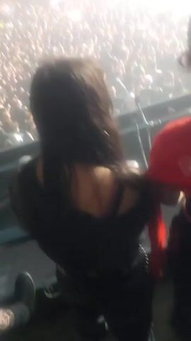 A person films a large crowd of people at a concert. The crowd is dense and energized, with many individuals holding up their phones. A bright spotlight illuminates the crowd, creating a foggy atmosphere. The camera is positioned from an elevated viewpoint, likely a balcony or platform. Some people in the crowd are raising their arms, and one person in the foreground makes an obscene gesture. The music playing has vocals, but its hard to discern.