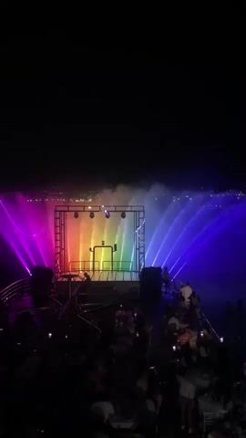 The video shows a nighttime outdoor event. In the center, a stage structure with lights is illuminated with shifting rainbow colors. Water fountains spray outwards from behind the stage, also lit with rainbow hues. A crowd of people is visible in the foreground, some seated at tables, others standing. Many are holding up their phones, presumably to record the event. The overall atmosphere appears to be a lively celebration or performance.
