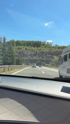 A car is driving on a highway towards a tunnel. The sky is blue with some clouds. The tunnel entrance is visible in the distance, illuminated by green lights. Several cars are driving on the highway, some ahead of the car from which the video is being filmed. The tunnel itself appears to be equipped with overhead lights that are also illuminated. The sides of the highway are lined with vegetation and rocky hills. The interior of the car is visible in the foreground, showing a dashboard and part of a seat.