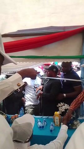The video shows a crowd of people gathered under a tent-like structure. A table covered in blue fabric is visible in the foreground, with water bottles, juice boxes, and flower arrangements on it. Several men stand around the table, shaking hands and interacting with each other. Some individuals are wearing hats, and the attire varies from casual t-shirts to more formal attire. A red, white, and green banner is draped overhead, adding to the festive or official atmosphere of the gathering. The overall impression is of a social or political event.