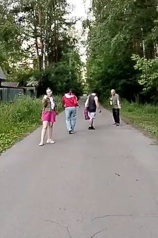 A group of four people walk down a paved path surrounded by trees. A woman in a pink skirt and a green jacket stands facing the camera. Behind her, three figures walk away from the camera. One figure in a red jacket walks ahead, followed by a person carrying a bag and another person holding a long object. The scene appears to be outdoors in a wooded area.