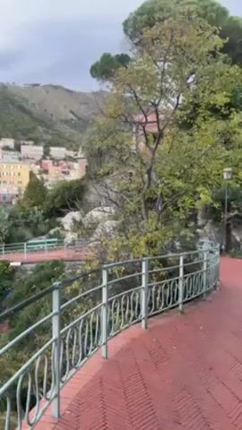 The camera pans slowly from right to left, revealing a town situated on a coast. The scene is shot from an elevated position along a walkway featuring a red herringbone-patterned brick surface and a light green railing. Trees and greenery line the side of the walkway. In the distance, a hillside densely populated with buildings painted in pastel shades of yellow, pink, and white is visible. Boats are docked along the water's edge. The sky is overcast, suggesting a cloudy day.