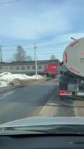 A vehicle is stopped on a road at a train crossing. A passenger train, with red and grey cars, is visible in the background, passing horizontally across the view. To the right of the frame is the back of a large, gray tanker truck. The road is wet, and piles of melting snow are visible on the side of the road. Overhead, power lines run parallel to the train tracks. The sky is a clear, light blue.  The camera appears to be mounted inside a car looking out over the hood.