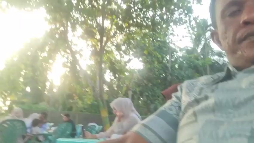 The video shows a group of people sitting at green tables and chairs outside in a shady, sandy area. Most of the women are wearing hijabs. Some are interacting with each other, while one woman is looking at her phone. Trees surround the area, and the sun is shining through the leaves. The camera is held by someone who is seated, with part of their arm visible in the foreground.