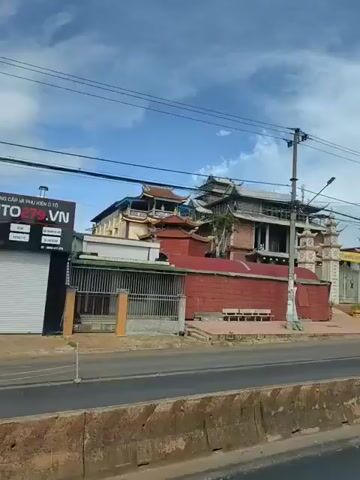 The video shows a street view of a building with traditional Asian architectural elements, possibly a temple or pagoda, under construction. The building features multiple levels with curved roofs and ornate decorations. A large entrance gate with decorative pillars is visible. A red brick wall surrounds parts of the property. Power lines crisscross the sky above. A person is riding a scooter past the building on the street in the foreground. The sky is cloudy.
