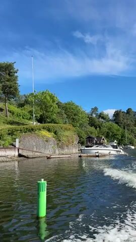 The video shows a boat moving through calm, green water. On the left bank, there is a small, modern white building with large windows, situated on a rocky slope covered with grass and trees. Further up the slope, a tall flagpole stands out against the blue sky. On the right bank, a larger, multi-level modern white house is visible, nestled among trees on a hill. Two white chairs are placed on the grass in front of this house. Another modern white house with red accents is also visible further up the hill. The water has white wakes from the boat, and a green buoy is visible in the foreground. The sky is clear and blue with a few wispy white clouds. The overall scene is one of serene natural beauty with modern architecture.