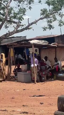 The video shows a scene of potential truck maintenance in what appears to be a village or roadside area. A group of people are gathered under a shaded structure, and a woman wearing a patterned yellow dress stands nearby. The focus then shifts to a stack of tires and a white truck parked nearby with its bed tilted upwards. A man in a purple robe stands near the truck's rear wheels, observing or working on it. An air compressor is positioned near the truck's front. In the background, there are other small structures and vehicles, suggesting a rural or less developed area. The ground appears to be mostly dirt or gravel.
