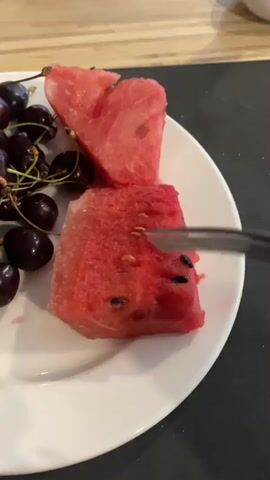 A close-up shot shows a white plate with several pieces of watermelon and a pile of dark cherries. One piece of watermelon is cut into a heart shape, and another is a rectangular slice. A fork is in the process of picking up a bite-sized piece of watermelon from the rectangular slice, which has several black seeds visible. The plate is on a dark surface, possibly a countertop.