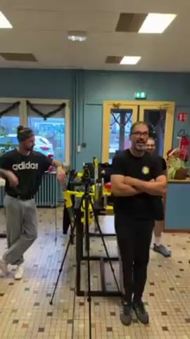 The video shows a group of people in what appears to be a training or recreational facility. One man in a black t-shirt and pants, with glasses, stands with his arms crossed, smiling and looking towards the camera. The group appears to be watching an arm wrestling match between two men at a table. The man on the left is wearing a dark t-shirt, while the man on the right wears a patterned dark green t-shirt. They are gripping each other's hands, their arms positioned on the table. The room has tiled floors and light blue walls with white ceilings. There are windows on one side and various fitness equipment visible in the background. The room seems to be set up for arm wrestling practice, as evident from the dedicated table and the 'Arm Fight Factory' sign on the wall.