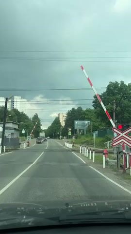 The video shows a view from the driver's seat of a car approaching a railroad crossing. The crossing gates are down, blocking the road, and a red light is illuminated on the crossing signal. The road is asphalt with white lane markings. On either side of the road are white posts with red tops. Trees and buildings are visible in the background. The sky is overcast with dark clouds. A few cars and people are visible on the road ahead, stopped at the crossing. The video captures the moment the gates begin to rise, allowing traffic to pass.