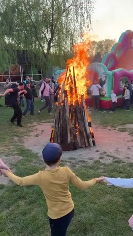 In a grassy outdoor area, a crowd of people is gathered around a large bonfire. Some people are holding hands and dancing in a circle around the fire. In the foreground, a boy in a yellow shirt and a girl in a white shirt and pink tutu are holding hands and dancing. In the background, there is a large inflatable play structure with bright colors. The bonfire is made of stacked logs and is burning brightly. The people in the crowd appear to be enjoying themselves. The video captures a lively and festive atmosphere.