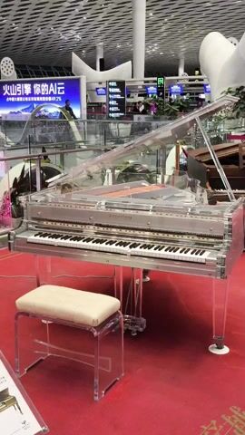 The video shows a Kay-erhing brand clear acrylic grand piano and matching bench situated on a red carpet inside a modern airport or convention center. The transparent design allows the inner workings of the piano to be visible. The bench has a white cushion. In the background are airport-style signs and advertisements in Chinese, as well as other pianos. The lighting is bright and artificial, highlighting the transparency of the piano and bench. There is no apparent action, interaction, or people within the main frame of the camera. The camera remains stationary, providing a full view of the piano.