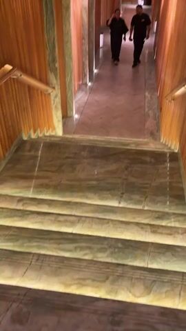 A first-person perspective video shows a downward view of marble stairs. Two men in dark uniforms are walking away from the camera down a hallway with wood-paneled walls and a textured metallic ceiling. The man closer to the camera is shorter and appears to be wearing a black t-shirt and black pants. The man further away is taller and wearing a black polo shirt with a logo on the left chest and black pants. The hallway is lit by recessed lights along the floor.