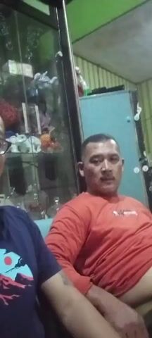 Two men are sitting indoors, possibly in a living room. The man on the left is wearing glasses and a dark blue t-shirt with a graphic design. He is smiling broadly and appears to be speaking. The man on the right is wearing a red t-shirt and is also smiling. Behind them, there is a glass-fronted cabinet filled with various decorative items, and a light blue cabinet or appliance. The lighting is dim, suggesting it is either nighttime or the room is not well-lit. The overall impression is of a casual and friendly interaction.
