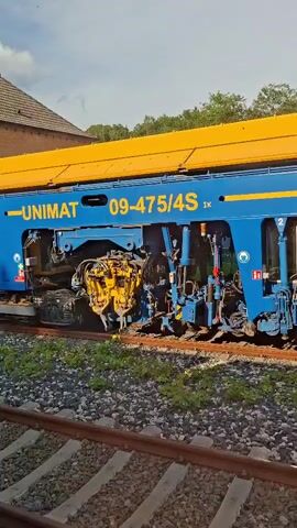 The video shows a close-up, side-view of a blue and yellow train car on railway tracks. The train car has the words "UNIMAT" and "09-475/4S" printed on its side. There are various mechanical components visible on the lower portion of the train car, including gears and hydraulic elements. The train car is stationary on a ballast track, with vegetation growing on the sides of the tracks. In the background, there are trees and a cloudy sky. The camera appears to be moving slightly, suggesting a panning shot.