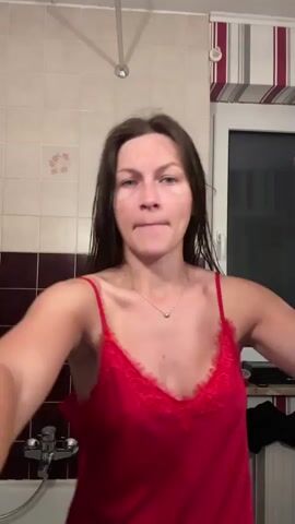 A young woman with long, wet, dark hair, wearing a red lace-trimmed camisole and a delicate silver necklace, stands in a bathroom and dries her hair. She holds a rose gold and white hairdryer in her left hand, moving it around her head, while her right hand gestures near her hair. She looks directly at the camera with varying facial expressions, including an open mouth and a slight pout. The bathroom features tiled walls with white and dark brown sections, a shower fixture, and a window with a striped curtain or blind visible in the background.