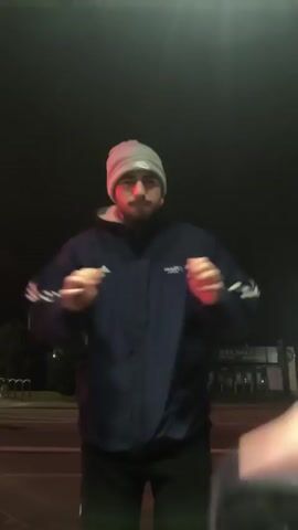 A man wearing a beanie, a dark blue jacket with white stripes on the sleeves, and black pants is standing outside at night. He is making boxing motions with his hands. There is a building with bright lights across the street and a street with bike racks on the left. The general scene is dimly lit. The man moves slightly during the video. Loud music plays in the background.