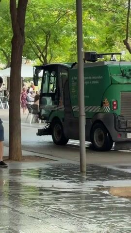 A man in a black t-shirt and denim shorts, carrying an orange shoulder bag, walks along a paved path. He passes a green street cleaner vehicle that is actively spraying water onto the ground. As the man walks through the sprayed water, his shorts get wet, and he looks down at them with a slight smile. The street cleaner, partially visible behind trees and a pole, has text on its side that reads "EL Prat" and "Netegem els contenidors amb aigua a pressió" (We clean the containers with pressurized water). Its license plate "E5900 BGZ" is visible on the rear. A black bicycle is parked near one of the trees. The scene is an outdoor urban environment, possibly a pedestrian area or sidewalk, lined with green trees. In the background, blurry figures and white tents suggest other activity or a market area. The time of day appears to be daytime.