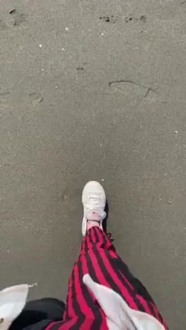 A person walks on a gray sand beach towards the water's edge. They are wearing white sneakers, and black pants with a red and white plaid shirt tied around their waist. The beach shows footprints in the sand.  Waves wash onto the shore. The water is a light blue-gray.  The sky is a pale overcast.