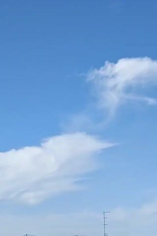 The video shows a bright blue sky with scattered white clouds. The clouds are wispy and appear to be moving slowly. In the lower part of the frame, a few buildings and a television antenna are visible. The overall scene is calm and peaceful, suggesting a pleasant day.