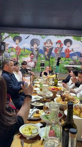 A well-lit indoor scene shows a large group of people sitting around a long wooden table laden with food and drinks, including wine bottles, carafes, salads, mashed potatoes, and a variety of other dishes. A man stands at the center of the table, facing the group, and appears to be speaking or performing. He holds a small object in his hand, potentially a lighter or a similar item. Several people are clapping along to music playing in the background, while another person is filming the scene with a smartphone. A colorful mural depicting historical figures in traditional attire is visible on the wall behind the group. The overall atmosphere seems celebratory and festive.