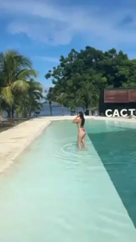 The video shows a woman in a red bikini standing in a shallow, turquoise-colored pool. She has her hands behind her head. The pool is surrounded by a light-colored concrete border and green grass, with palm trees and other foliage in the background. Beyond the trees, there is a view of the ocean and a clear blue sky with some clouds. To the right of the pool, there's a wooden building with the word "CACTUS" displayed on it. The camera remains static throughout the video.
