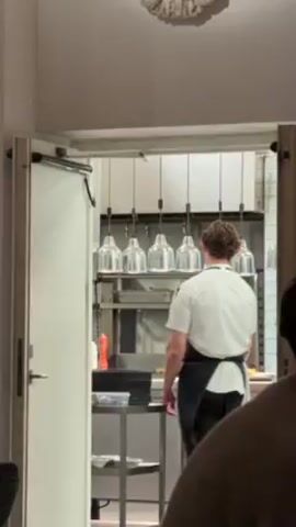 The video shows a kitchen setting from the vantage point of someone sitting in a dining area. Two chefs, one male with short brown hair wearing a white chef's coat and a black apron, and a female with red hair pulled back wearing a similar uniform, are seen preparing food. The man places a platter on the counter, and the woman picks it up and exits the kitchen to presumably serve the dish. Above the counter are several hanging pendant lights. There is a stack of plates on the counter, a metal shelving unit holding various items, and a partial view of another diner in the foreground. The kitchen is visible through an open doorway with a white frame. There is ornate decoration on the ceiling in the dining area.