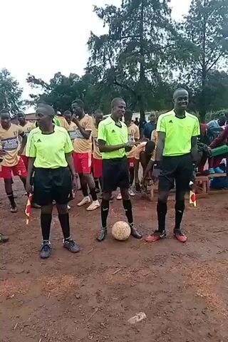 A group of people is standing on a dirt field. Three people in neon green shirts and black shorts are in the center. One is a woman. They appear to be officials or referees for a sports event. A soccer ball sits on the ground near their feet. Behind them are other people, some wearing athletic jerseys. The people are all African or of African descent. Tall trees are in the background. The sky is overcast.