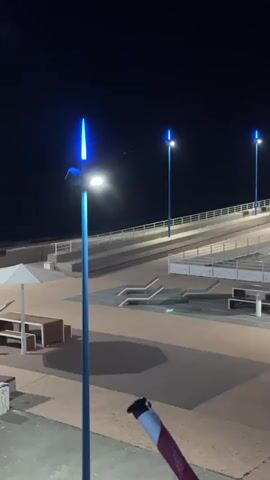 A video taken at night shows an outdoor area with several streetlights. The streetlights have blue lights at the top and white lights at the side. There are also benches and a ramp visible in the background. The camera is pointed downwards at a slight angle, with a hand holding what appears to be a vape pen in the foreground, partially obscuring the view. The area is dimly lit by the streetlights, with shadows cast on the ground.