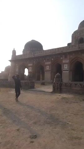 The video shows a large building made of stone or brick, possibly a mosque, with a large dome on top. There are two men walking in front of the building, and a woman in a hijab is looking at the ruins in the background. The ground is dry and dusty.
