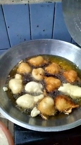 A shallow metal pan filled with hot oil is seen from a high angle. Several pieces of dough are being fried in the oil. The dough pieces are irregular in shape and range in color from pale white to golden brown. They are bubbling and some are partially submerged in the oil. A slotted metal spoon or skimmer is being used to stir or move the dough pieces within the pan. The pan is on a surface that appears to be a tiled countertop or table. The overall scene suggests food preparation, likely in a kitchen. The image is well-lit, and there are no indications of time of day or weather. The location context is given as Rambipuji, Indonesia.