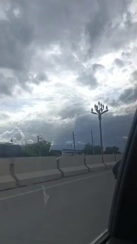 A video shows a street scene with a cloudy sky. A concrete barrier runs along the side of the road. On the other side of the barrier, there are utility poles and streetlights. In the distance, mountains can be seen. The video is filmed from inside a vehicle, as indicated by the presence of a car's side mirror in the lower right corner.