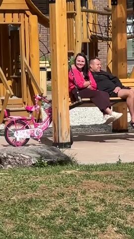 The video shows a wooden playground swing with a bench seat. A man and a woman are sitting on the swing. The man is on the right, wearing a black t-shirt and black shorts. He is wearing flip-flops. The woman is on the left, wearing a pink jacket over a dark top and a dark skirt. She has her arm around the man. A pink bicycle is parked next to the swing. The playground is made of wood and has a climbing net. In the background, there is a building with yellow and beige sections. The ground is a mix of grass and a paved surface. The scene appears to be taking place in daylight.