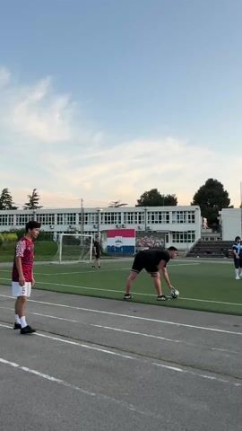 The video shows an outdoor soccer field with several people in athletic wear.  Some people are walking around, and one person is picking up a soccer ball.  The sky is blue with some clouds, and the sun is setting in the background.  There is a building behind the soccer field with windows and a Croatian flag.  There are also some light poles around the field.  Someone throws the ball and the other people play.