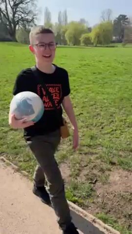 A young, light-skinned person with short hair and glasses, wearing a black t-shirt and khaki pants, is walking along a path in what appears to be a park or playground. They are carrying a white and blue soccer ball. The person is smiling and looking towards the camera at times. The background includes green grass, trees, and wooden fencing. In one shot, a red structure is visible in the distance. They walk past the fencing, presumably towards the playground or park area. They have a black strap or bag slung over their shoulder. The lighting suggests a bright, sunny day.