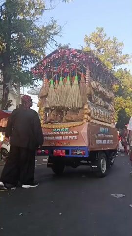 A group of people, some dressed in traditional clothing, are seen standing near a decorated truck. The truck is adorned with brooms and what appear to be sacks or bundles. It is parked on a street lined with trees and buildings. The sky is clear and blue. One person is walking away from the camera, while others are standing and looking towards the truck. There is a scooter parked on the left side of the image. The overall scene suggests a parade or a cultural event.