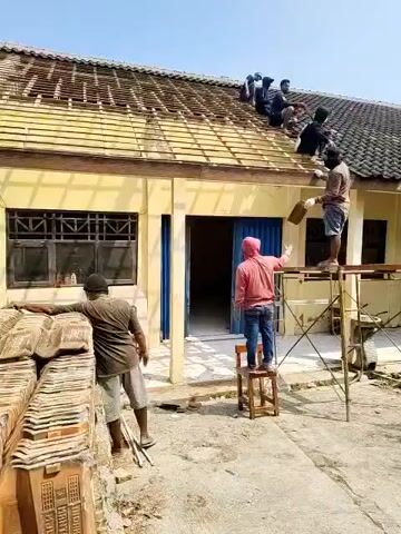 Six men are working on a yellow, one-story building with a tiled roof. The roof is in disrepair, with some tiles missing and others exposed. The men are on the roof, near the edge, and on scaffolding, laying or repairing tiles. One man is on a stepladder, reaching up to hand a tool to a man on the roof. Another man is standing on a wooden chair and holding out his hand to receive something from the man on the stepladder. A man in a brown shirt and shorts is leaning against a stack of tiles on the ground. The sky is clear and blue. The ground is dirt and some concrete. There is scaffolding to the right of the building.