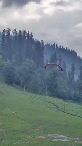 A paraglider is shown in mid-air against a backdrop of a forested mountain. The paraglider, which is red and orange, is suspended above a green, grassy slope. Below the paraglider, several people are visible on the slope, some walking and others standing. The mountain behind the slope is covered in dense trees, and the sky above is overcast with clouds. The scene appears to be a popular spot for outdoor activities.