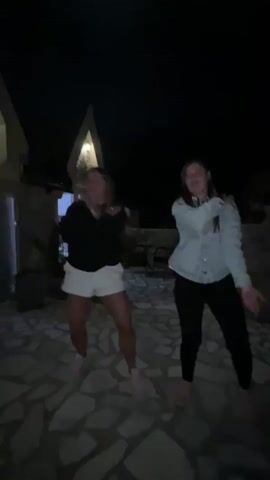 Two young women are dancing outdoors at night on a stone-paved patio or terrace in Sotonići, Montenegro. The setting is dimly lit by artificial lights, illuminating the ground and parts of the surrounding buildings, one of which has a distinctive pointed architectural feature. In the background, there's some outdoor furniture including chairs and a table.

Both women are performing synchronized dance moves, primarily involving arm swings and hip swaying. The woman on the left is wearing a black long-sleeved top, white shorts, and light-colored sneakers. The woman on the right is dressed in a light blue denim-style jacket, black pants, and light-colored shoes, possibly espadrilles. They appear to be enjoying themselves, smiling and laughing throughout the performance. At one point, the woman on the left turns her back to the camera, briefly touching her backside as part of the dance, while the woman on the right continues to face forward with similar movements.