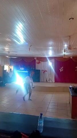 A person in traditional African attire is dancing on a tiled floor in a banquet hall. The individual is wearing a white lace top, a striped skirt, and a headdress. The dancer twirls, kneels, and uses a feather duster as part of their performance. In the background, there's a stage decorated with red and white curtains.  A DJ setup with flashing blue lights and a sound system is on the stage, along with several people behind the DJ equipment. The hall is lit by overhead fluorescent lights and ceiling fans. A water bottle is placed on a table in the foreground. Several people are seated in the background, and the video is taken from the front of the stage.