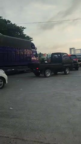 The video shows a view from a paved area looking towards a gas station. A black pickup truck is parked in the foreground, facing right. Behind it, a dark-colored SUV is also parked, and further back, there are other vehicles and buildings. To the left, a large purple truck is parked. The sky is overcast, and the lighting suggests it is either early morning or late afternoon.