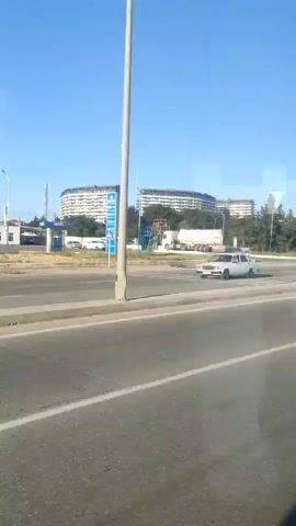 The video shows a street scene with a blue sky. On the left, there is a gas station sign. In the center, there are several large trucks on a road. In the background, there are buildings and trees. There is also a pedestrian crossing sign visible. The camera appears to be moving, possibly from a vehicle.