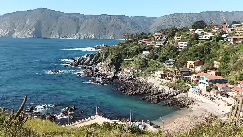 The video shows a coastal scene with a bay surrounded by rocky cliffs and hills. The water is turquoise near the shore and deeper blue further out.  There are some houses built along the cliffs, overlooking the bay. A small beach can be seen at the base of the cliffs. In the distance, large hills stretch along the coastline. The foreground shows vegetation, including grasses and cacti. The camera pans slowly, showing a wider view of the bay and the distant hills.